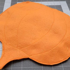 May include: Orange felt pumpkin with stitched lines, resembling a pumpkin's shape. The pumpkin has a stem and is ready for crafting.