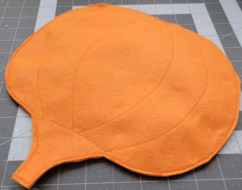 May include: Orange felt pumpkin with stitched lines, resembling a pumpkin's shape. The pumpkin has a stem and is ready for crafting.