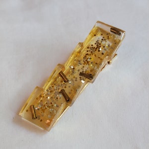 May include: A rectangular hair clip made of clear resin with gold glitter and small gold-colored cylindrical beads. The clip has a gold-colored metal clasp.