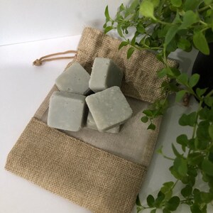 May include: A burlap bag with a drawstring closure contains several small, gray, square-shaped soap bars. The bag is sitting on a white surface next to a green plant.