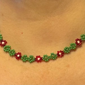 May include: A green and red beaded necklace with small flower designs. The necklace is made of tiny beads and features a repeating pattern of green leaves and red flowers with white centers.
