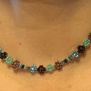 May include: Beaded necklace featuring a floral design. The necklace has small, round beads in shades of brown, black, light blue, and teal. The beads are strung together to create a delicate, eye-catching accessory.