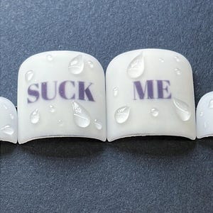 Op de afbeelding: Witte nail art met de woorden "SUCK ME" in paarse letters. De nagels hebben een glanzende afwerking met waterdruppel details. De achtergrond is donkergrijs.
