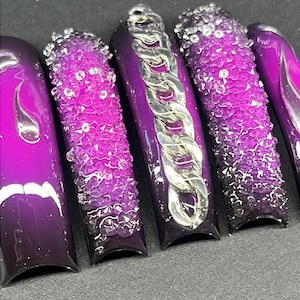 May include: Close-up of five long, purple artificial nails. The nails feature a gradient effect, transitioning from a deep purple at the base to a lighter shade at the tip. Some nails have a textured, crystal-like design, while one features a silver chain.