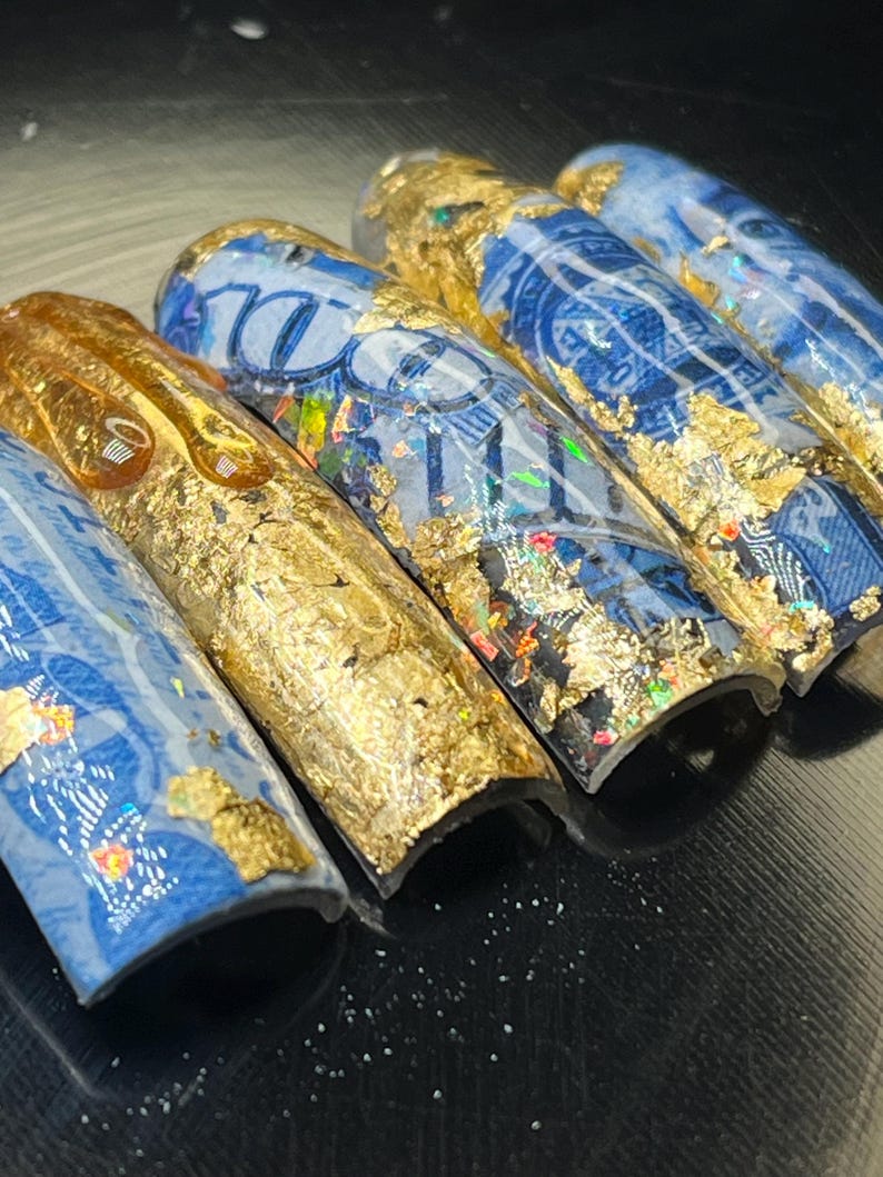 May include: Five blue and gold nail tips with a dollar bill design. The tips are curved and have a glossy finish.