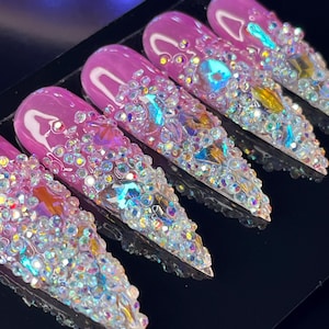 May include: Five pink stiletto-shaped false nails with iridescent rhinestones covering the entire nail surface.