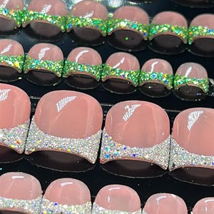 Puede incluir: Una colección de uñas postizas para los pies de varios tamaños. Las uñas presentan una base rosa claro con un diseño brillante plateado o verde en la punta. Las uñas están dispuestas sobre una superficie negra.