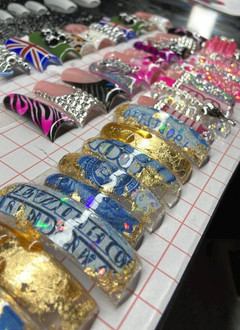 May include: A set of false nails with various designs, including a zebra print, a Union Jack flag, and a design featuring gold leaf and a US dollar bill.