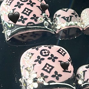 Könnte beinhalten: Nahaufnahme von rosa Nagelkunst mit schwarzen Blumen- und Logomustern. Die Nägel sind mit schwarzen Herzakzenten und funkelnden Strasssteinen verziert. Das Design ist vor einem dunklen Hintergrund platziert.