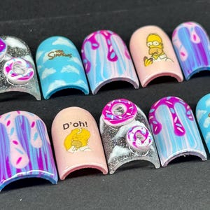 Puede incluir: Un conjunto de uñas postizas con diseños inspirados en Los Simpson. Las uñas muestran varios diseños, incluyendo a Homer Simpson, donuts y el logo del programa, con colores vibrantes como rosa, azul y morado. Algunas uñas tienen un acabado brillante.