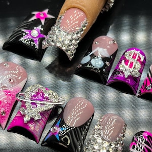 May include: A set of ten false fingernails with various designs. The designs include a star, a heart with a planet, a dollar sign, and a zebra pattern. The nails are decorated with rhinestones.