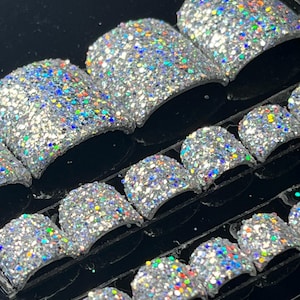 May include: A set of silver glitter press-on nails in various sizes. The nails are arranged in rows on a black background.