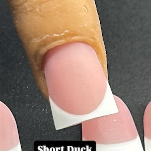 May include: Close-up of a finger with a Short Duck nail design. The nail is a soft pink with a white tip. The text "Short Duck" is in a black rectangle.