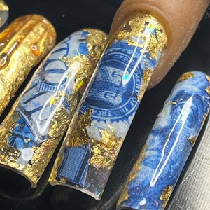May include: Close-up of several long, blue and gold nail tips with a design of US currency and gold leaf. The nail tips are on a black surface.