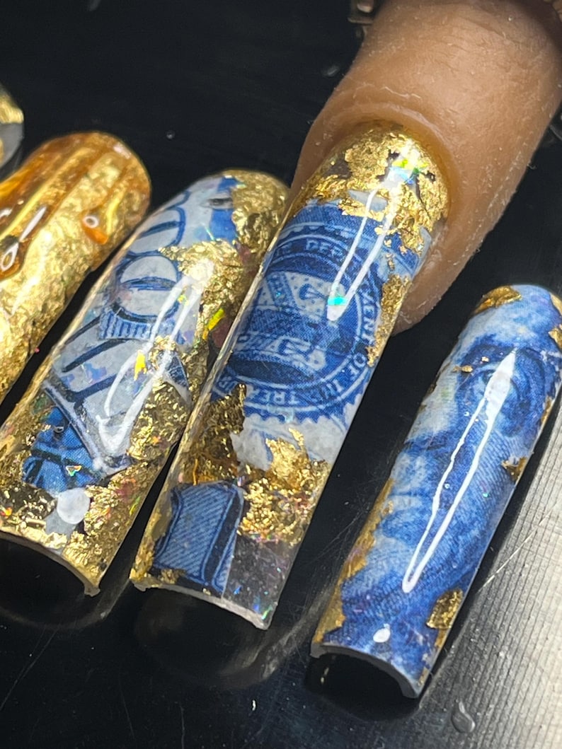 May include: Close-up of several long, blue and gold nail tips with a design of US currency and gold leaf. The nail tips are on a black surface.