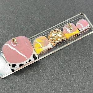 Puede incluir: Un conjunto de cinco uñas artificiales con varios diseños. Una uña presenta una base rosa con un patrón blanco y negro. Otra tiene un diseño de mármol amarillo y blanco. La uña central está cubierta de pedrería dorada. Las otras dos uñas tienen diseños rosas y amarillos.