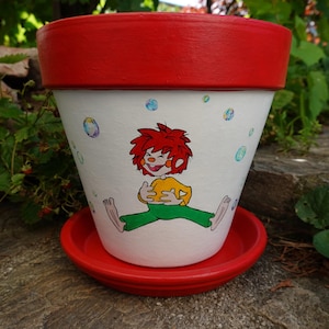 Könnte beinhalten: Ein weißer Terrakotta-Blumentopf mit einem leuchtend roten Rand und passendem Untersetzer. Auf der Vorderseite befindet sich eine handgemalte Illustration einer Figur mit stacheligen roten Haaren, gelbem Oberteil und grüner Hose, umgeben von Blasen.