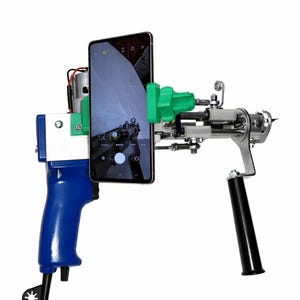 May include: A handheld tufting gun with a blue handle, white and silver components, and a black grip. A smartphone is attached to the gun with a green mount. The phone displays a camera interface.