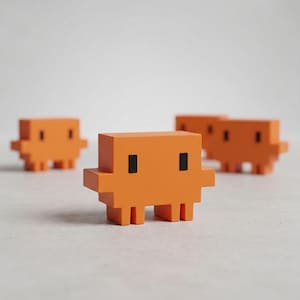Può includere: Quattro figurine arancioni pixelate a forma di creatura, con occhi neri rettangolari e piccole zampe. Sono disposte su una superficie grigio chiaro, una a fuoco e le altre sfocate sullo sfondo. Il design ricorda i personaggi dei videogiochi retrò.