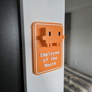 May include: An orange "Employee of the Month" sign with a pixelated character design. The sign is mounted on a white wall. The text is white and the border is white.