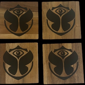 May include: Set of four square wooden coasters. Each coaster features a dark, stylized eye and floral design. The coasters are made of light brown wood with a visible grain pattern, and the design is in a dark color.