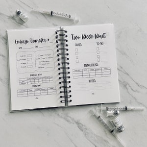 Puede incluir: Una agenda en blanco y negro con el texto "Embryo Transfer #" y "Two Week Wait" en la portada. La agenda tiene una encuadernaci&oacute;n en espiral e incluye secciones para llevar un registro de los medicamentos, las metas y las tareas pendientes.