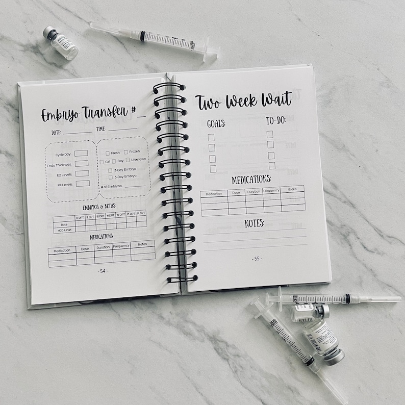 Puede incluir: Una agenda en blanco y negro con el texto "Embryo Transfer #" y "Two Week Wait" en la portada. La agenda tiene una encuadernaci&oacute;n en espiral e incluye secciones para llevar un registro de los medicamentos, las metas y las tareas pendientes.