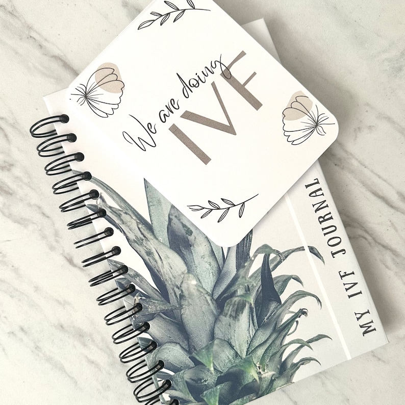 Puede incluir: Un diario blanco con una encuadernaci&oacute;n en espiral negra y un dise&ntilde;o de pi&ntilde;a en la portada. El diario tiene el texto "My IVF Journal" impreso en la portada. Una tarjeta blanca separada con el texto "We are doing IVF" y un dise&ntilde;o de mariposa se coloca encima del diario.