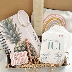 Puede incluir: Una caja de regalo llena de artículos, incluyendo un diario con un diseño de piña, un pastillero blanco con los días de la semana, una bolsa de arcoíris, un juego de tarjetas con el texto "We are doing IUI", un bolígrafo de oro rosa con un adorno de piña y una pegatina que dice "Strong, brave, beautiful, warrior".