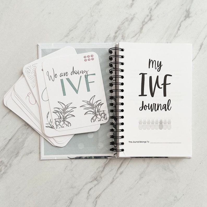 Puede incluir: Un diario blanco con el texto "my IVF journal" y una fila de ilustraciones de pi&ntilde;a. El diario tiene una encuadernaci&oacute;n en espiral negra y una tarjeta blanca con el texto "We are doing IVF" y dos ilustraciones de pi&ntilde;a.