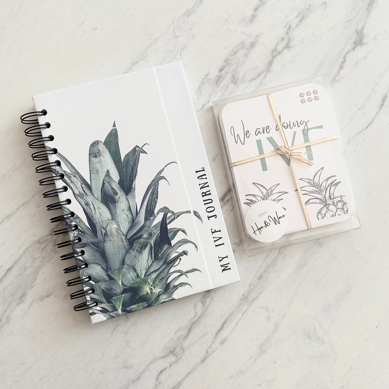 Puede incluir: Una libreta blanca de espiral con el texto "My IVF Journal" en la portada. La portada presenta una ilustraci&oacute;n de pi&ntilde;a verde y blanca. Tambi&eacute;n se muestra una peque&ntilde;a tarjeta con el texto "We are doing IVF".