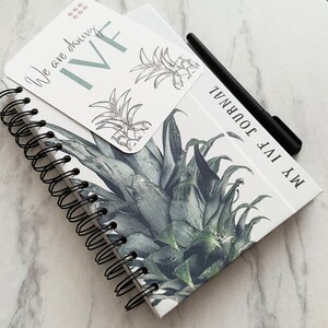 Puede incluir: Un cuaderno blanco de espiral con un dise&ntilde;o de pi&ntilde;a y el texto "My IVF Journal" en la portada. Una peque&ntilde;a tarjeta con el texto "We are doing IVF" est&aacute; unida a la parte delantera del cuaderno.