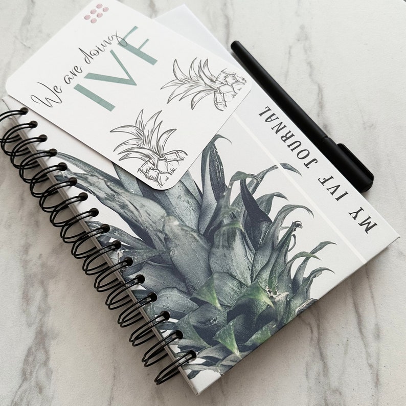 Puede incluir: Un cuaderno blanco de espiral con un dise&ntilde;o de pi&ntilde;a y el texto "My IVF Journal" en la portada. Una peque&ntilde;a tarjeta con el texto "We are doing IVF" est&aacute; unida a la parte delantera del cuaderno.