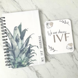 Puede incluir: Un diario blanco con un dise&ntilde;o de pi&ntilde;a y el texto "My IVF Journal" en la portada. Una tarjeta blanca con el texto "We are doing IVF" y dise&ntilde;os florales est&aacute; al lado del diario.
