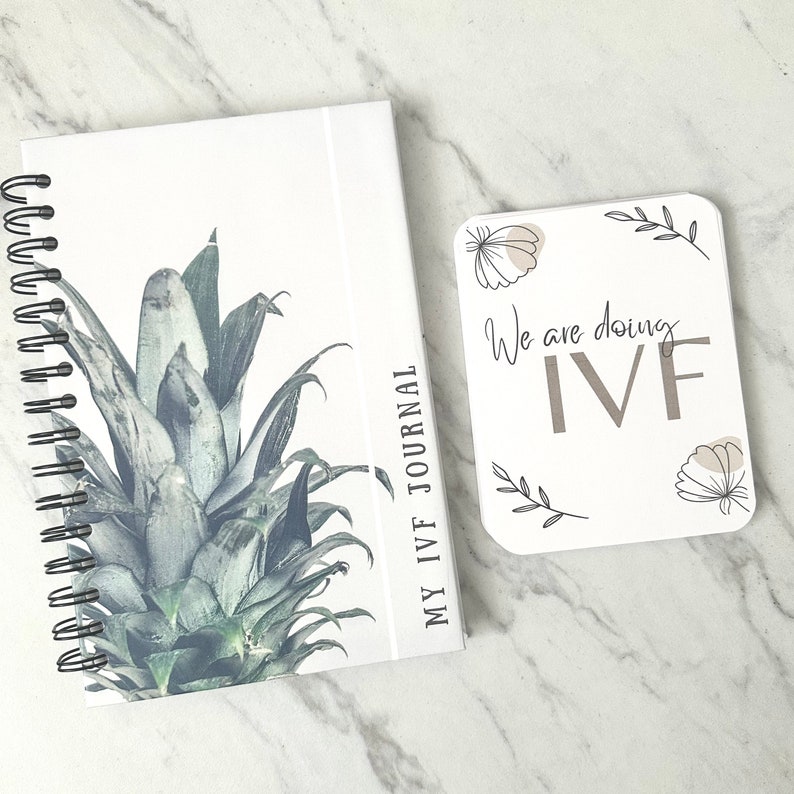 Puede incluir: Un diario blanco con un dise&ntilde;o de pi&ntilde;a y el texto "My IVF Journal" en la portada. Una tarjeta blanca con el texto "We are doing IVF" y dise&ntilde;os florales est&aacute; al lado del diario.