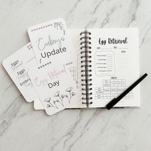 Puede incluir: Una libreta blanca con una encuadernaci&oacute;n en espiral negra. La libreta tiene el t&iacute;tulo "Recuperaci&oacute;n de &oacute;vulos" e incluye secciones para la fecha, la hora, las notas y una tabla para hacer un seguimiento del desarrollo de los embriones. La libreta est&aacute; abierta en una p&aacute;gina con un bol&iacute;grafo negro encima.