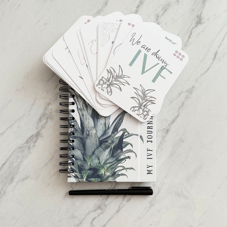 Puede incluir: Un diario blanco con un dise&ntilde;o de pi&ntilde;a y el texto "My IVF Journal". Una pila de tarjetas blancas con el texto "We are doing IVF" e ilustraciones de pi&ntilde;as est&aacute;n encima del diario. Un bol&iacute;grafo negro est&aacute; al lado del diario.