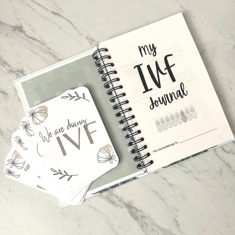 Puede incluir: Un diario blanco con el texto "my IVF journal" en la portada. El diario tiene una encuadernaci&oacute;n en espiral y est&aacute; abierto en una p&aacute;gina en blanco con el texto "This journal belongs to..." en la parte inferior. Una pila de tarjetas blancas con el texto "We are doing IVF" y dise&ntilde;os florales est&aacute; en el fondo.