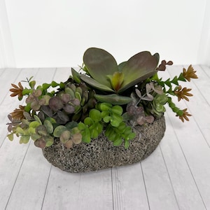 May include: An artificial succulent arrangement featuring a variety of green, brown, and purple-toned plants. The foliage is clustered together and set into a textured, oval-shaped gray stone-like planter with a porous, rough surface finish.