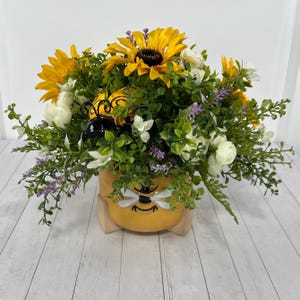 May include: A yellow ceramic planter shaped like a bee face with black antennae, filled with an arrangement of artificial yellow sunflowers, white roses, small green leaves, and sprigs of purple flowers. The planter has a smooth, rounded base.