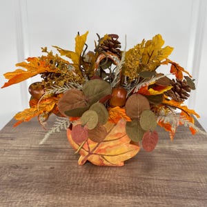 May include: A fall-themed centerpiece featuring a leaf-shaped container filled with faux autumn foliage. The arrangement includes orange, yellow, and brown leaves, pinecones, and berries. The container is painted with fall colors.