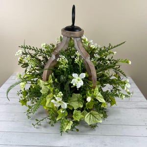 May include: A decorative wooden lantern filled with an arrangement of greenery and white flowers. The lantern has a rustic, weathered appearance with a black metal handle. The arrangement includes various types of green foliage and delicate white blossoms.