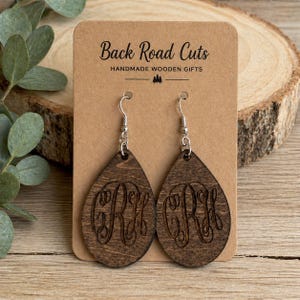 May include: A pair of teardrop-shaped wooden earrings with a dark brown finish. Each earring features a carved monogram design and silver-colored ear wires. The earrings are displayed on a brown card with the text "Back Road Cuts".