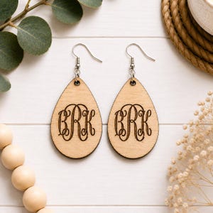 May include: A pair of teardrop-shaped wooden earrings featuring a laser-engraved monogram with the letters BRK in a decorative script font. The earrings are suspended from silver-toned metal fishhook wires for pierced ears.