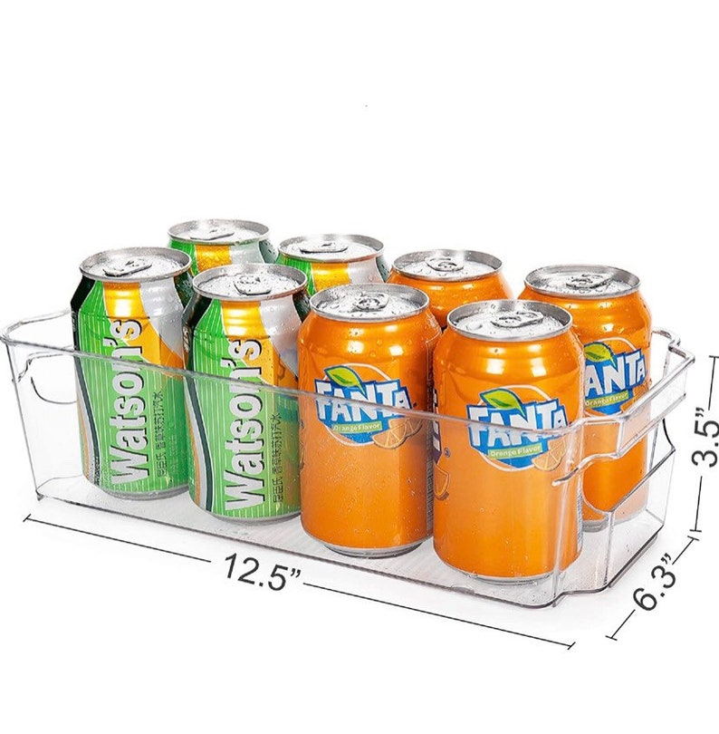 May include: A clear plastic refrigerator organizer with a handle, holding six cans of soda. The cans are a mix of green and orange, with the brands Watson's and Fanta. The organizer measures 12.5 inches wide, 6.3 inches deep, and 3.5 inches high.