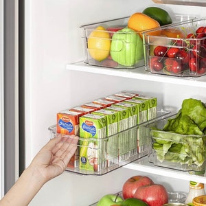 May include: Clear plastic storage containers in a refrigerator, filled with fruit, vegetables, and juice boxes. The containers are stacked on shelves and labeled "Juver Disfruta".