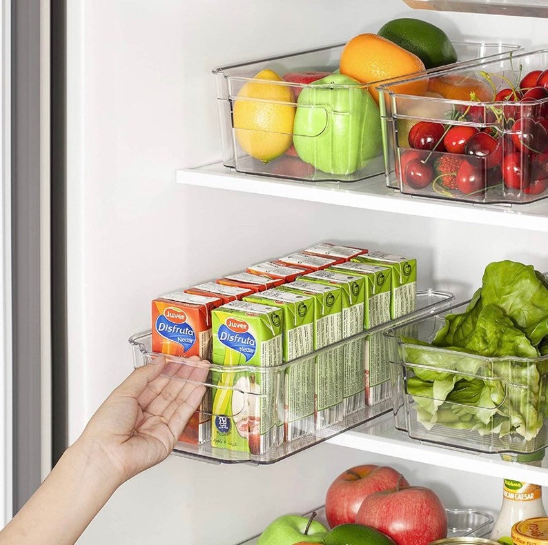 May include: Clear plastic storage containers in a refrigerator, filled with fruit, vegetables, and juice boxes. The containers are stacked on shelves and labeled "Juver Disfruta".