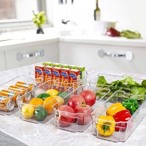 May include: Clear plastic refrigerator storage bins with dividers, filled with various fruits, vegetables, and juice boxes. The bins are labeled "Disfruta" and are stacked on top of each other.