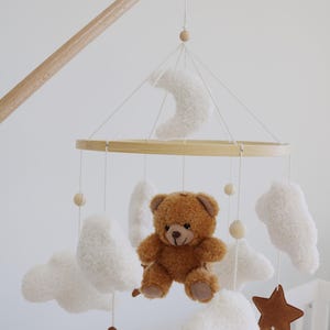 May include: A baby mobile featuring a brown teddy bear, white clouds, brown stars, and a white crescent moon. The mobile is suspended from a wooden arm and ring, creating a whimsical nursery decoration.