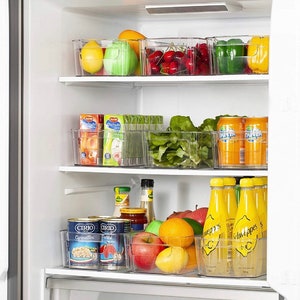May include: A refrigerator with clear plastic storage containers filled with various fruits, vegetables, and beverages. The containers are stacked on three shelves. The top shelf contains red cherries, green bell peppers, and yellow bell peppers. The middle shelf contains green lettuce, orange juice boxes, and cans of orange soda. The bottom shelf contains canned goods, oranges, lemons, apples, and bottles of yellow lemonade.